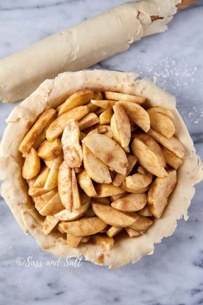 A top view of a pie crust filled with sliced apples seasoned with cinnamon. A rolling pin with dough is in the background on a marble surface.