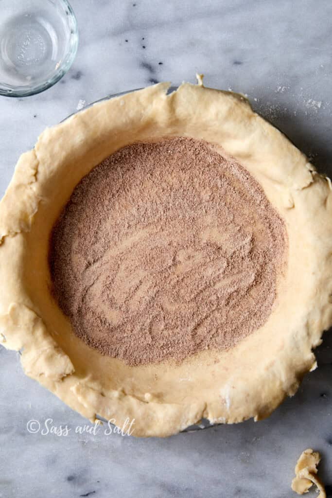 A round pie crust filled with a mixture of cinnamon, flour and sugar, displayed on a marble countertop. A small glass bowl is partially visible in the top-left corner.