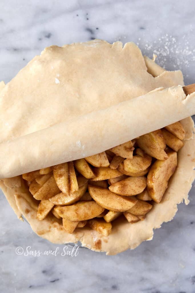 A partially assembled apple pie with spiced apple slices on a dough base, and a rolling pin rolling out the top crust, placed on a marble surface.