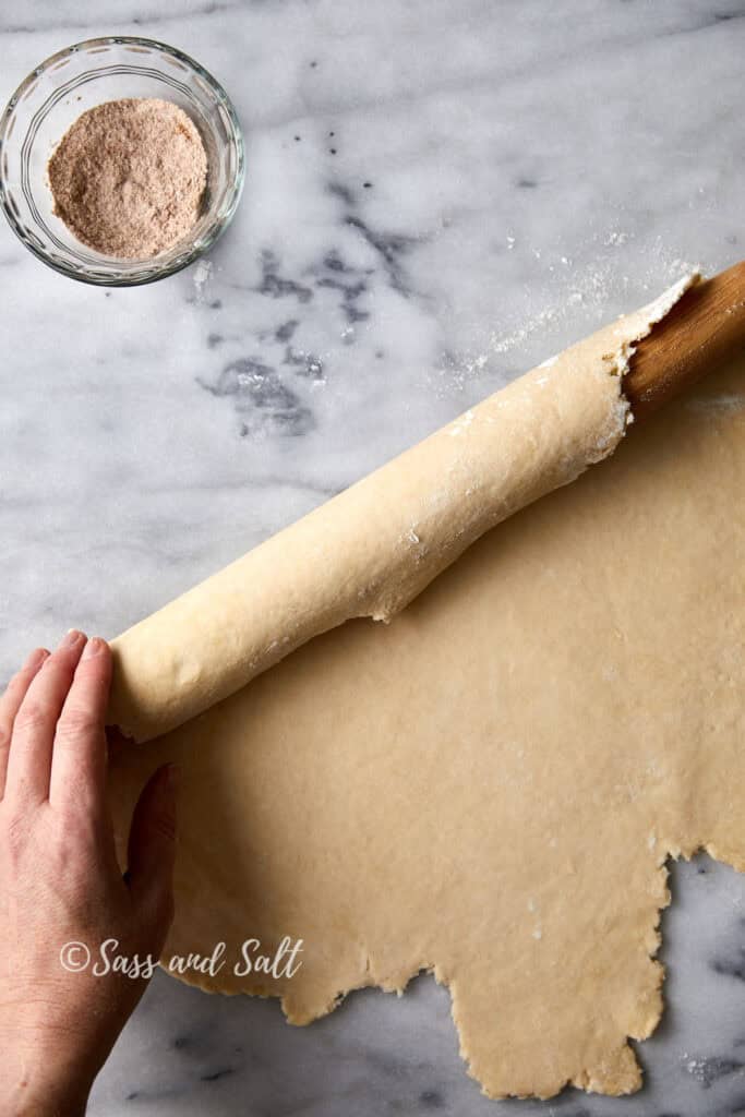 A hand rolls out pie dough with a wooden rolling pin on a marble surface. A small glass bowl with a brown spice mixture is nearby. The dough is partially rolled up, ready for baking.