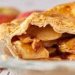 Close-up of a freshly baked apple pie with a golden, all-butter pie crust, revealing juicy cinnamon-coated apple slices spilling out. Blurred apples are visible in the background.