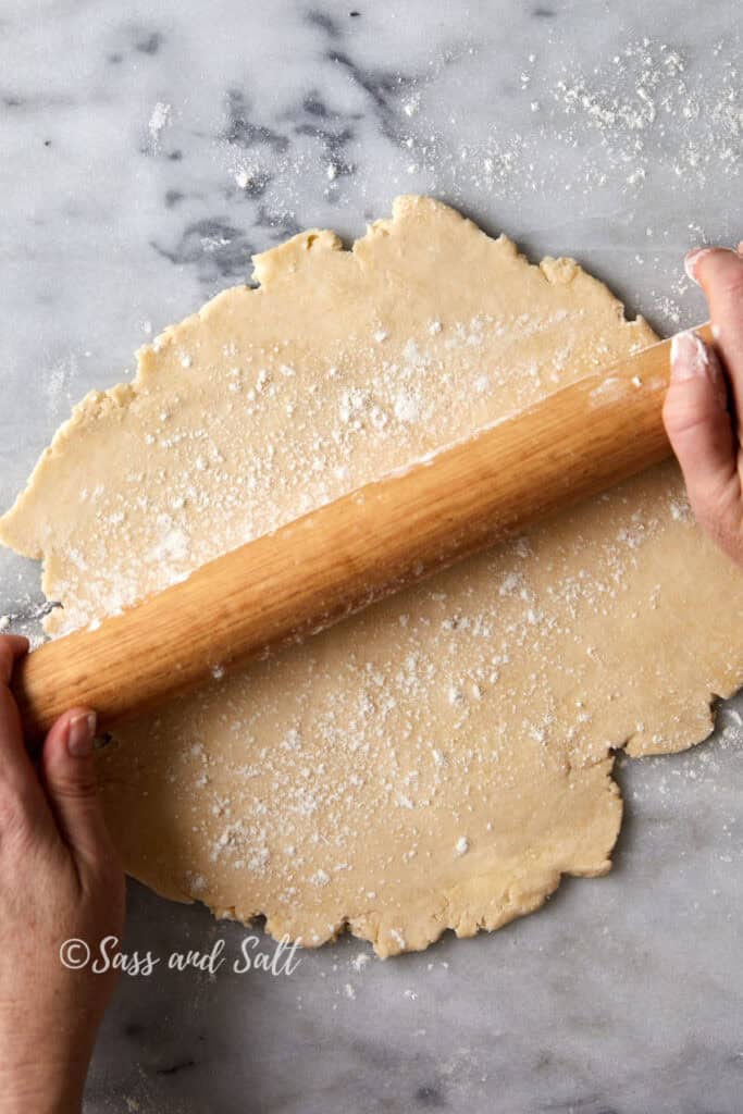 Hands using a wooden rolling pin to flatten dough on a floured marble surface. The dough is lightly dusted with flour.