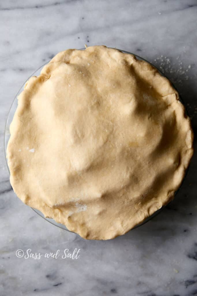 A pie with an uncooked dough crust covers a round pie dish, set on a marble countertop. The dough appears slightly textured and is evenly spread over the dish. The image is watermarked with the text "Sass and Salt.