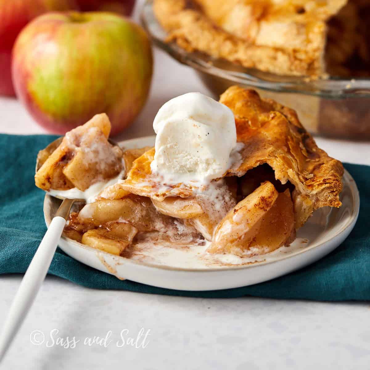 A slice of apple pie with a golden crust sits on a plate, topped with a scoop of melting vanilla ice cream. A white fork rests beside the pie, and fresh apples are in the background. The pie dish is partially visible.