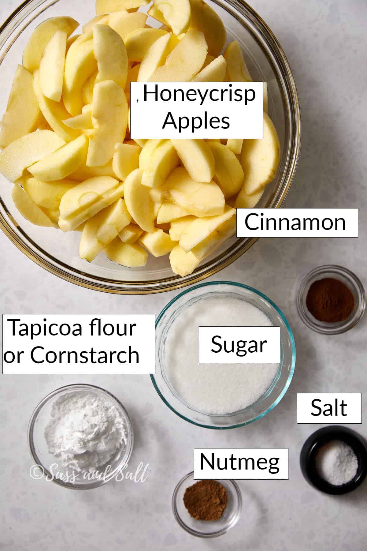 Top view of ingredients for an apple pie on a countertop. A bowl of sliced Honeycrisp apples, small bowls of cinnamon, sugar, nutmeg, salt, and tapioca flour or cornstarch are labeled and arranged around the apples.
