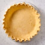 A photo of an unbaked pie crust with crimped edges in a glass pie dish, sitting on a light-colored, speckled countertop. The crust is evenly rolled out and pressed into the dish, ready for filling and baking.
