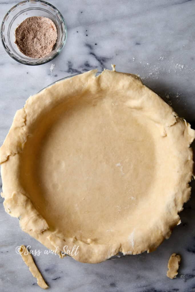 A pie crust is neatly placed in a pie dish on a marble surface, ready for filling. A small glass bowl with a mixture of cinnamon, flour and sugar sits nearby. Dustings of flour can be seen on the marble background.