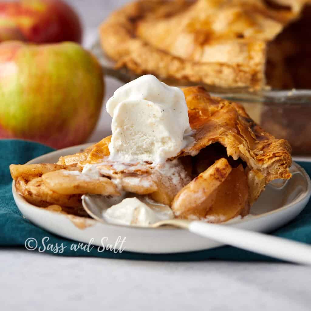 A slice of apple pie topped with a scoop of vanilla ice cream on a white plate, with a fork beside it. The background includes a whole pie in a dish and two fresh apples, with a teal cloth beneath the plate.