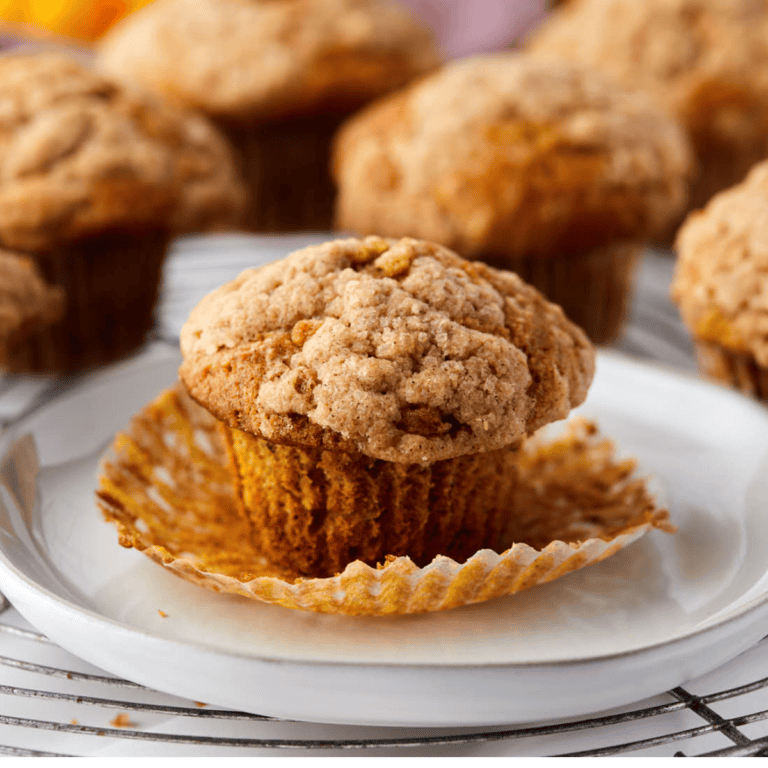 A close-up of a pumpkin banana muffin on a white plate, its wrapper partially peeled back to reveal its moist, crumbly texture. Several similar muffins are blurred in the background, all resting on a cooling rack.