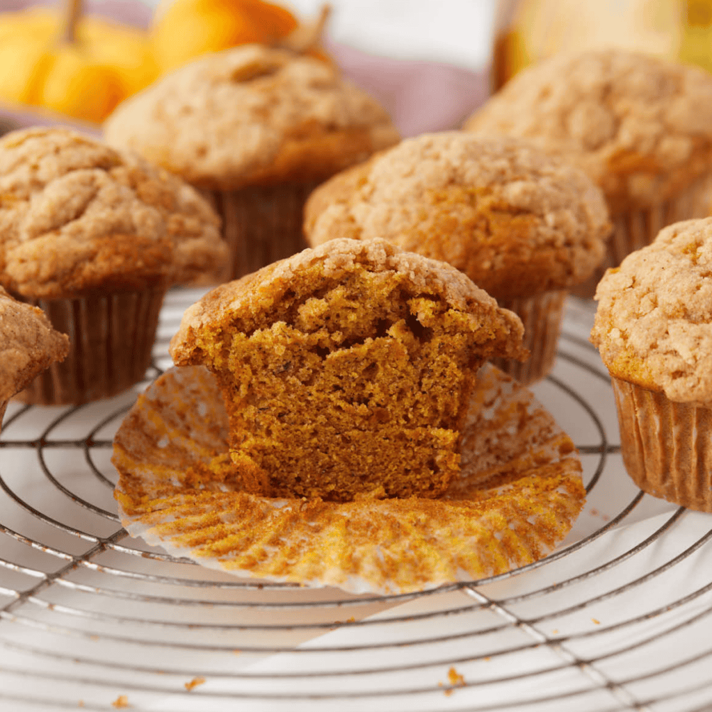 A close-up of freshly baked pumpkin banana muffins on a cooling rack. One muffin is cut open, revealing its moist, spiced interior—an easy fall recipe. Mini pumpkins and a yellow cloth are blurred in the background.