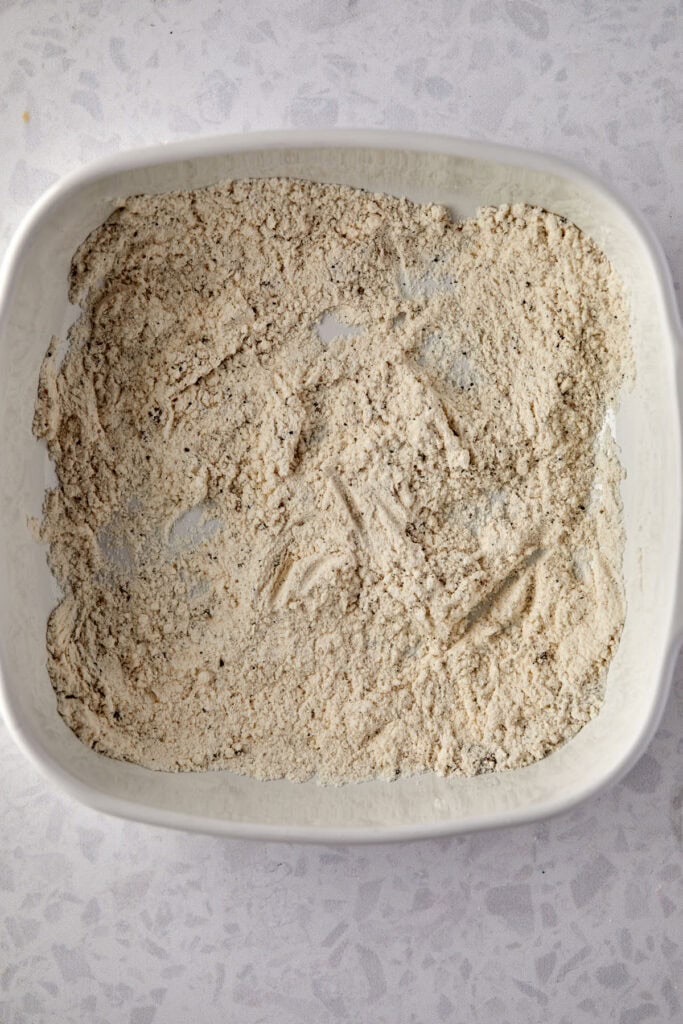 A white rectangular baking dish filled with flour and spices, on a light gray speckled countertop.