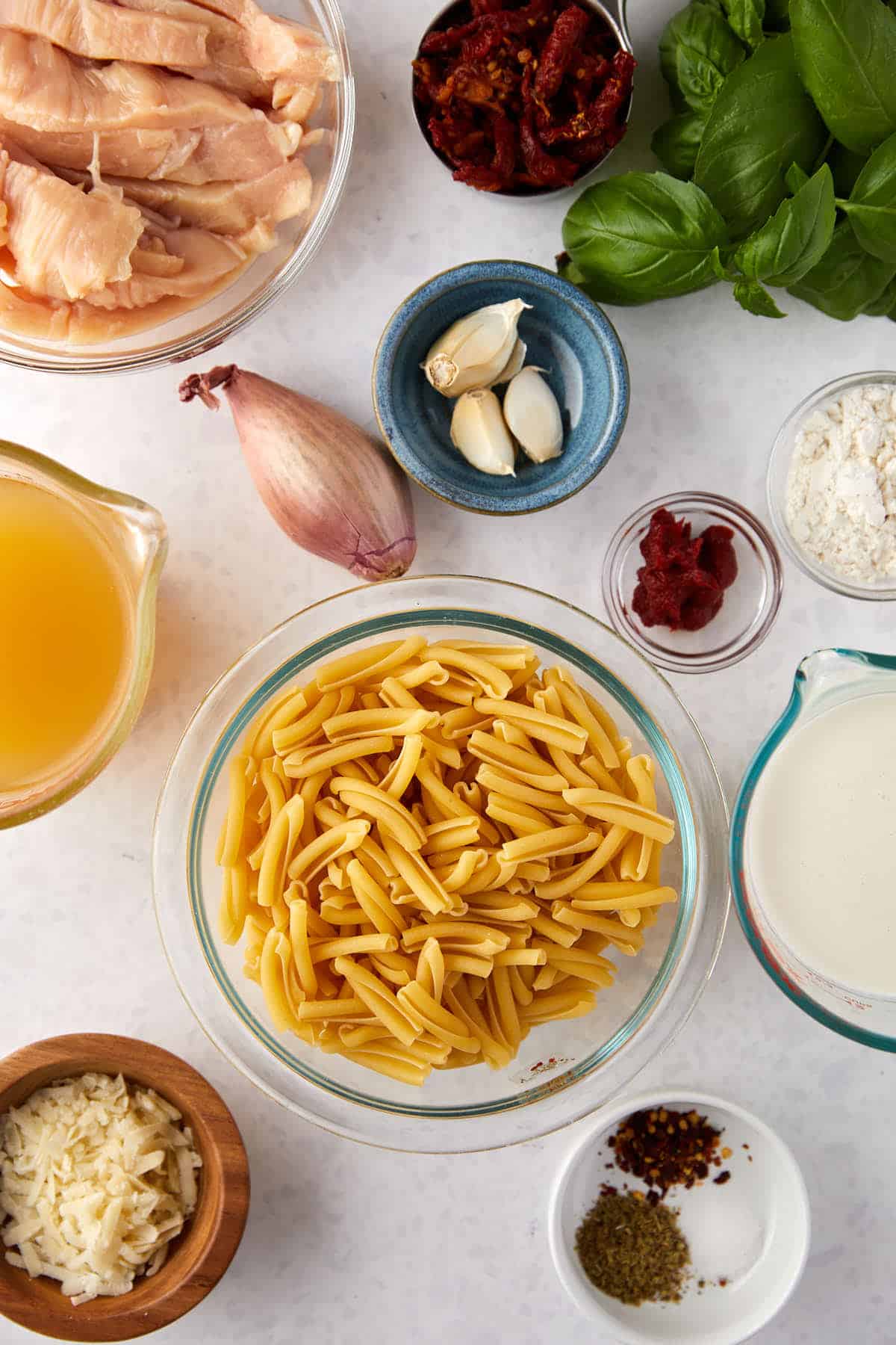 Top view of cooking ingredients on a white surface, including raw chicken, dried pasta, garlic cloves, onion, fresh basil, sun-dried tomatoes, cheese, spices, and liquids in bowls and containers, arranged neatly for preparation.