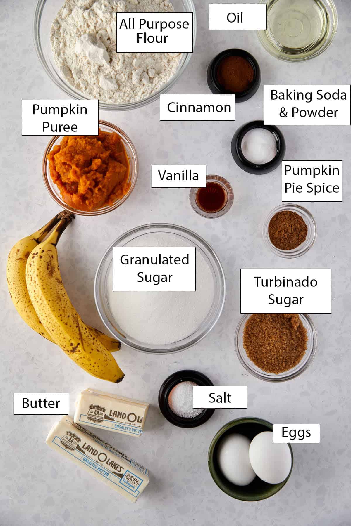 Ingredients for moist and delicious pumpkin banana muffins are arranged on a surface, including all-purpose flour, oil, pumpkin puree, cinnamon, baking soda, pumpkin pie spice, bananas, sugar, turbinado sugar, butter, salt, vanilla eggs - perfect for easy fall recipes like Pumpkin Banana Muffins.