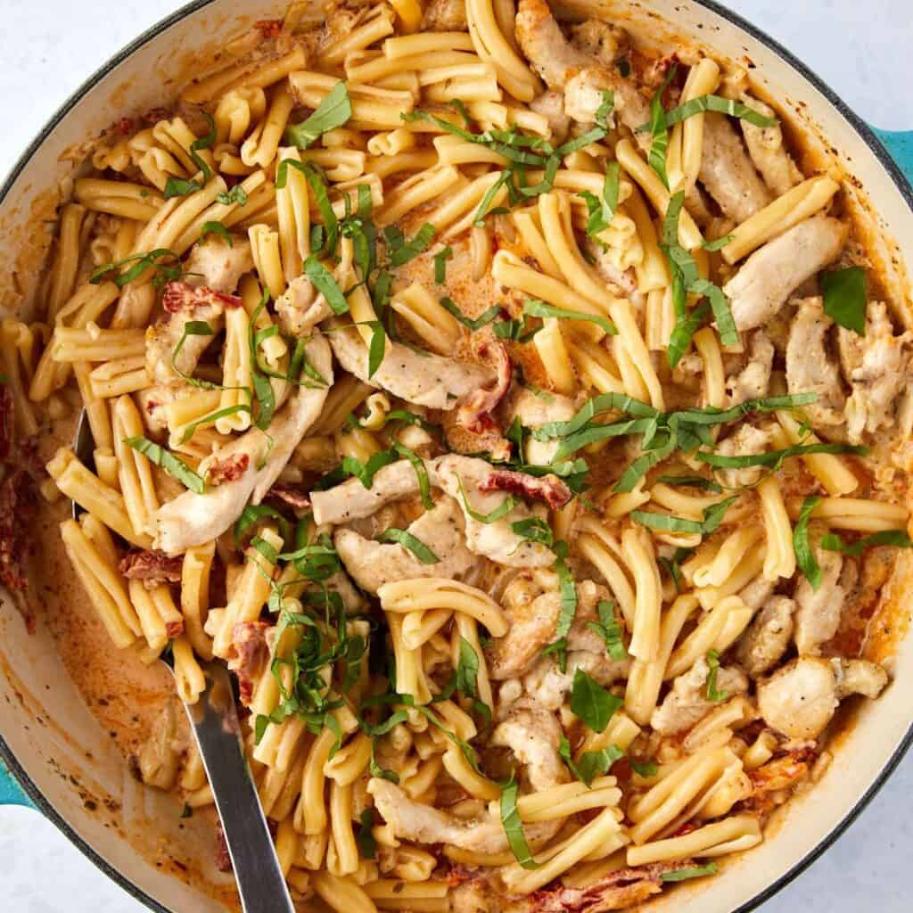 A skillet filled with creamy marry me chicken and pasta, sliced chicken, and sun-dried tomatoes, topped with freshly chopped basil. A large spoon rests in the pan, ready for serving.
