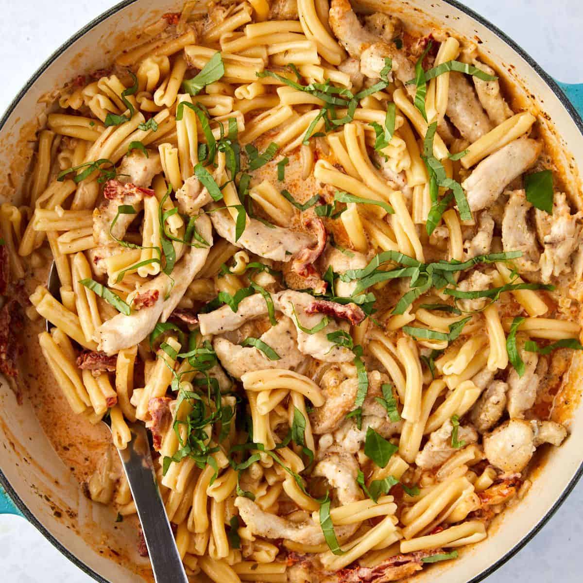 A skillet filled with creamy marry me chicken and pasta, sliced chicken, and sun-dried tomatoes, topped with freshly chopped basil. A large spoon rests in the pan, ready for serving.