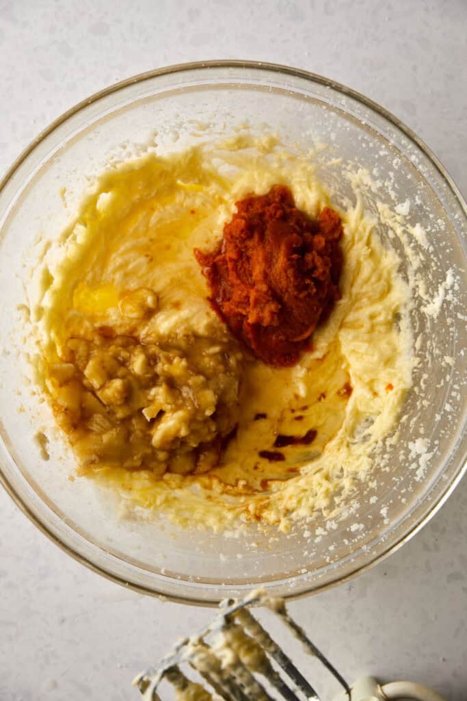 A glass mixing bowl with mashed bananas, pumpkin puree, and oil added to butter, sugar and egg mixture.  A whisk attachment from an electric mixer is partially visible at the bottom.