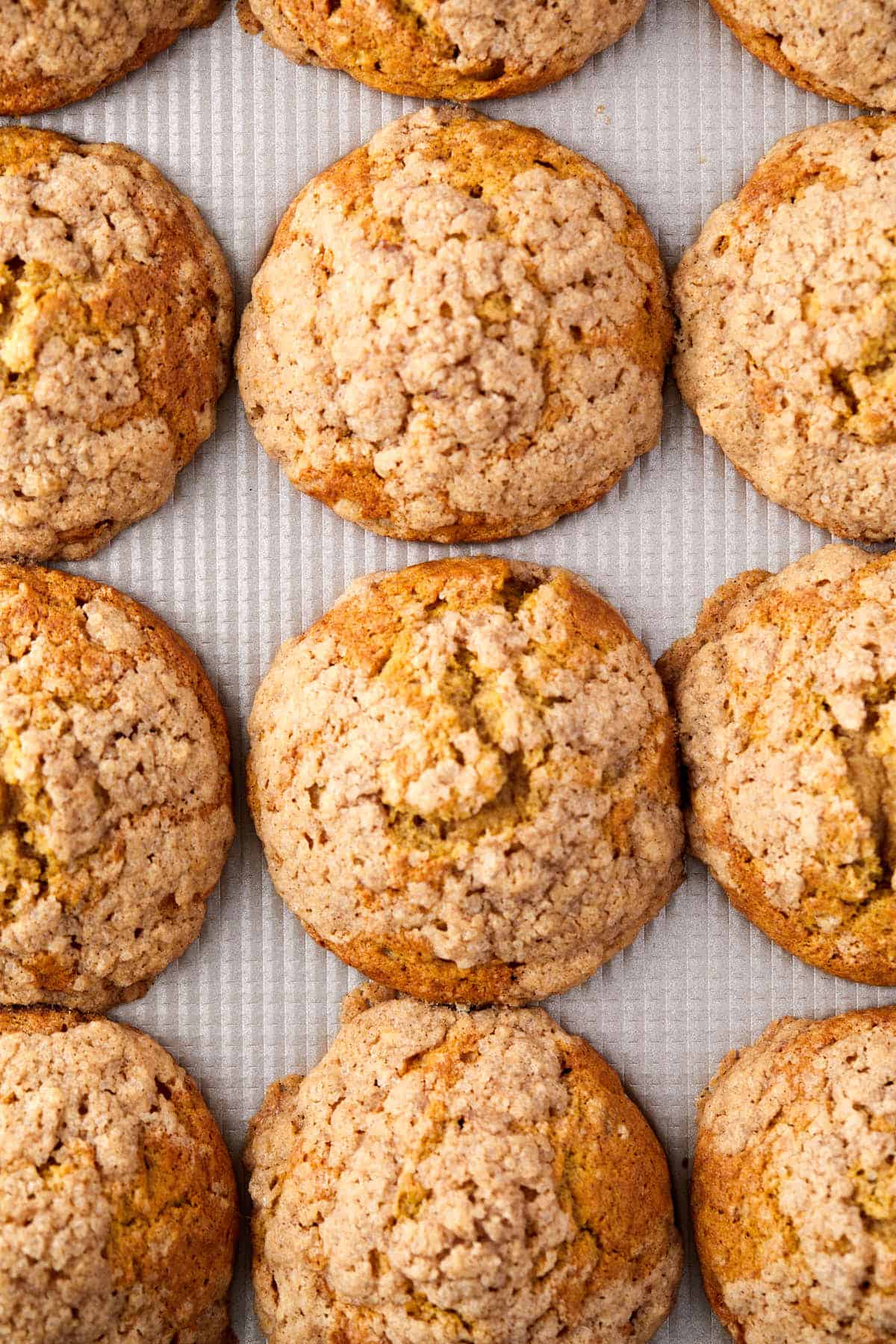 Rows of freshly baked, golden-brown cookies with a crumbly texture are arranged closely together on a ribbed baking sheet. The cookies have a slightly cracked surface, suggesting a soft interior.