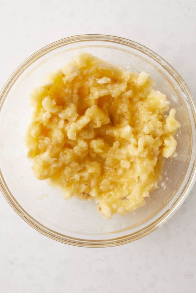 A glass bowl filled with mashed banana on a light-colored surface. The banana is a soft, chunky texture and light yellow in color.