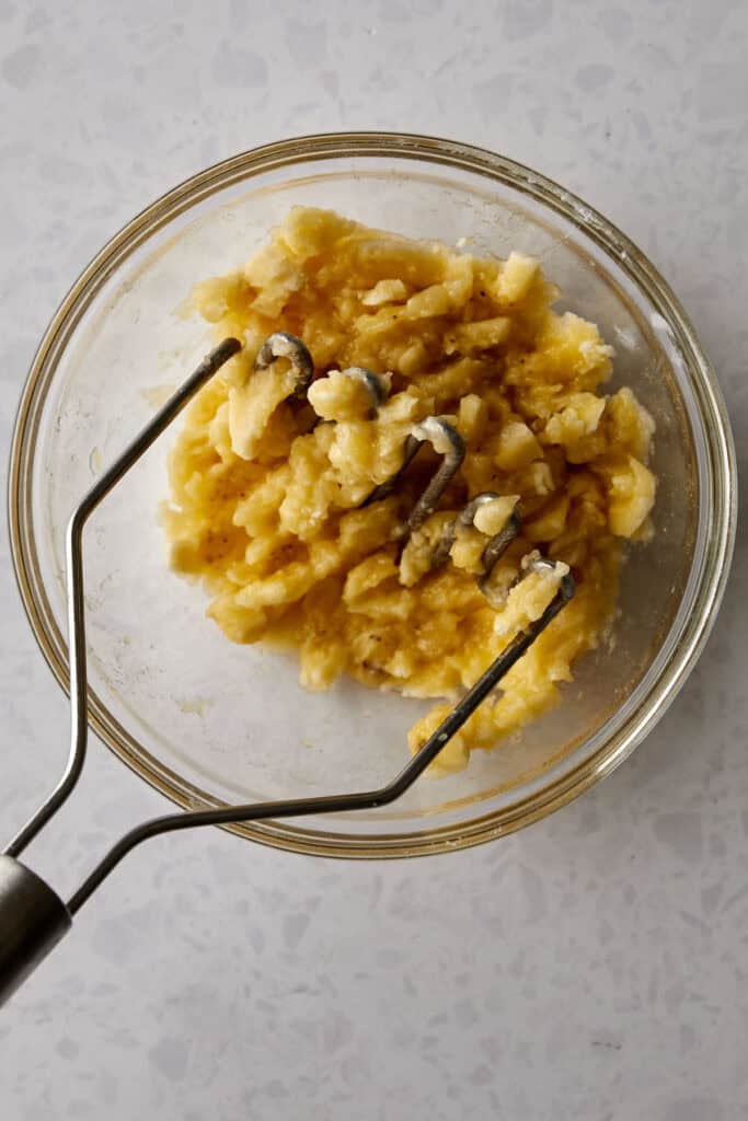 A glass bowl containing mashed bananas with a metal masher resting inside. The bowl is placed on a light gray countertop.