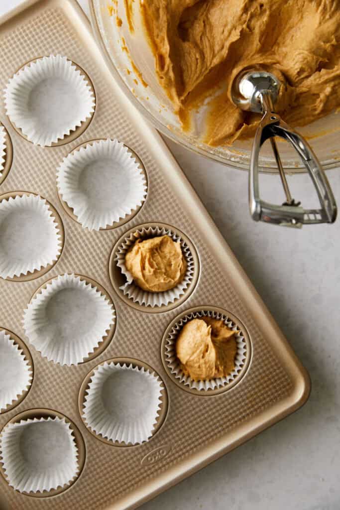 Muffin pan with paper liners partially filled with brown batter from a mixing bowl and ice cream scoop on a marble countertop.