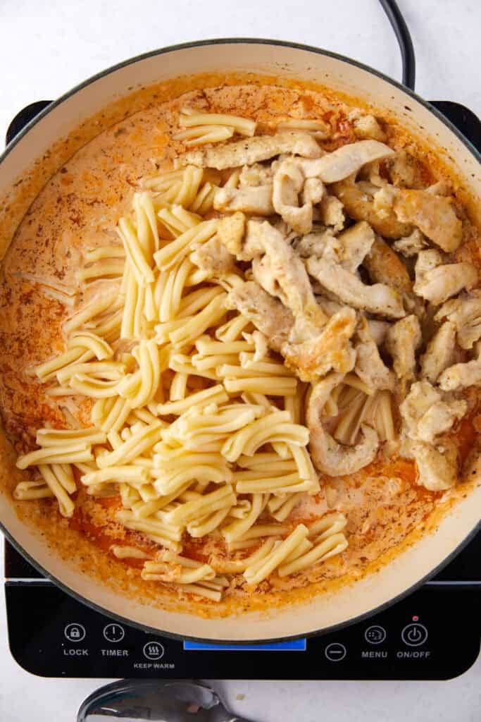 A pot on a stove contains pasta and sliced chicken mixed in a creamy, orange sauce. The pasta and chicken are partially combined, showing both ingredients clearly. The stove display is visible on the front.