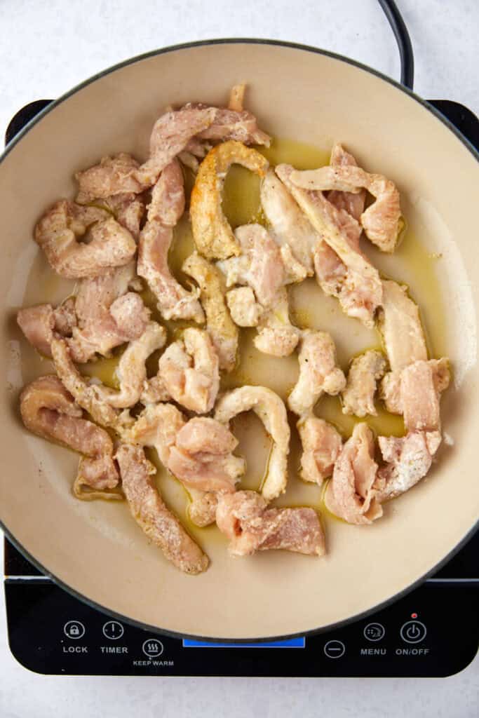 Raw chicken strips cooking in a pan with olive oil on a stovetop. The meat is beginning to brown and is evenly distributed across the surface of the pan. The stovetop controls are visible at the bottom.