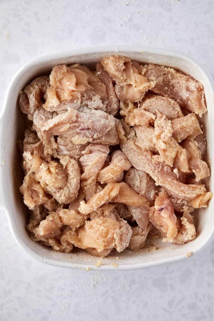 A white ceramic bowl filled with raw, seasoned chicken pieces. The meat is coated with a light layer of flour  ready for cooking. The background is a light-colored surface.