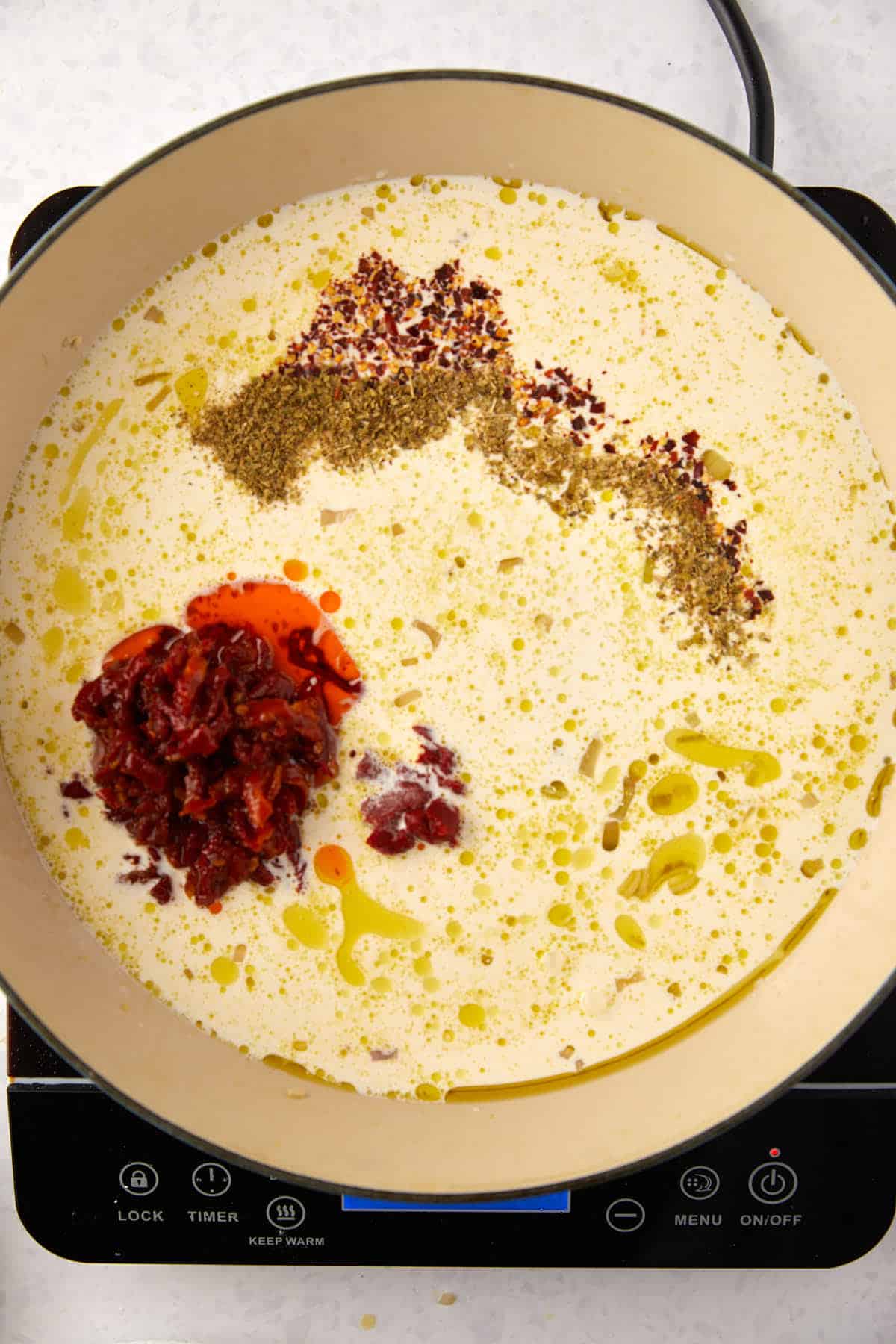 A pot on an electric stove containing cream, olive oil, sun dried tomatoes, and spices. The cream is mixed with seasoning and garnished with olive oil droplets, ready for cooking.