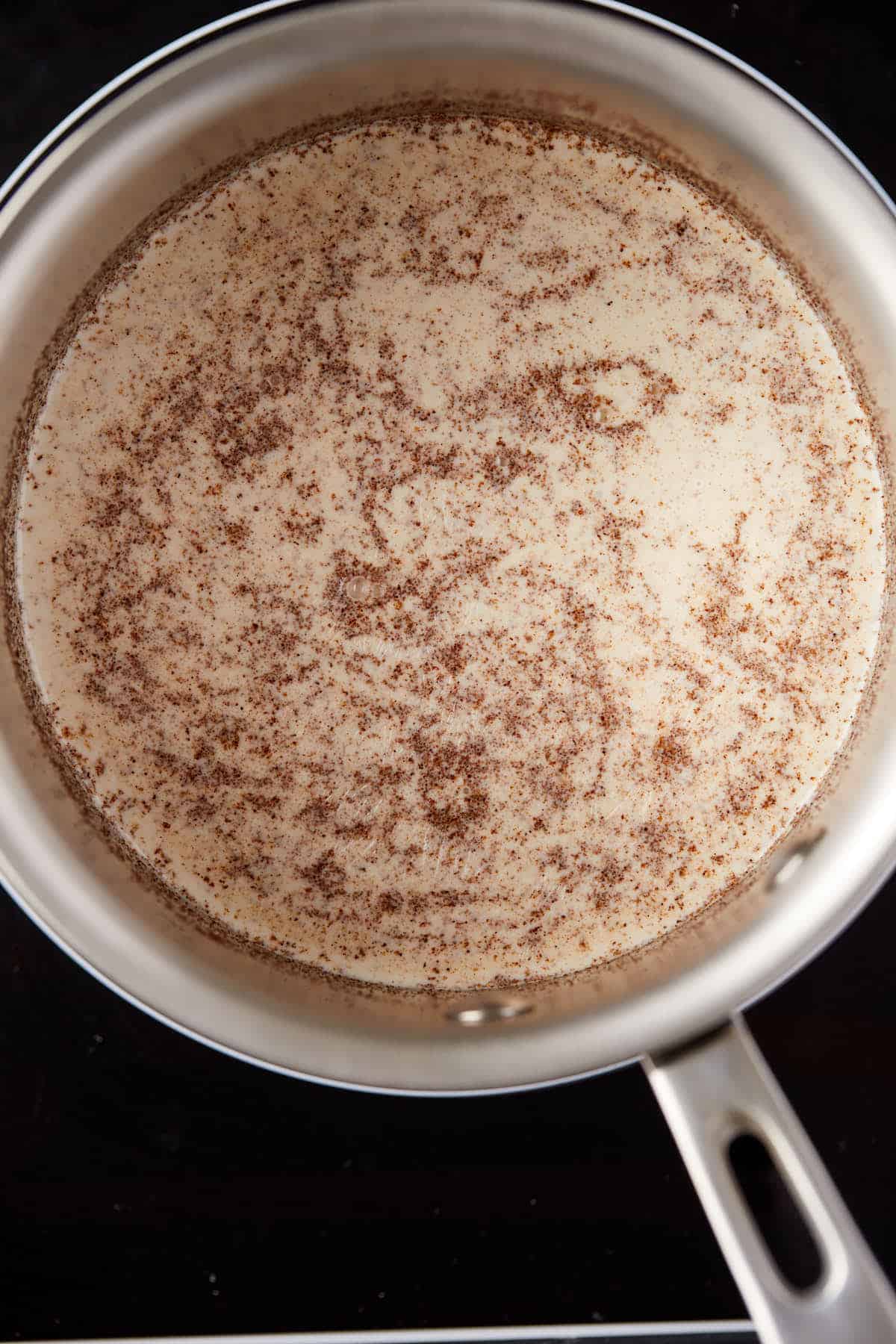 A saucepan on a stove containing a simmering mixture of milk with specks of cinnamon and spices visible on the surface. The handle of the saucepan extends outward, showing a bit of the stovetop underneath.