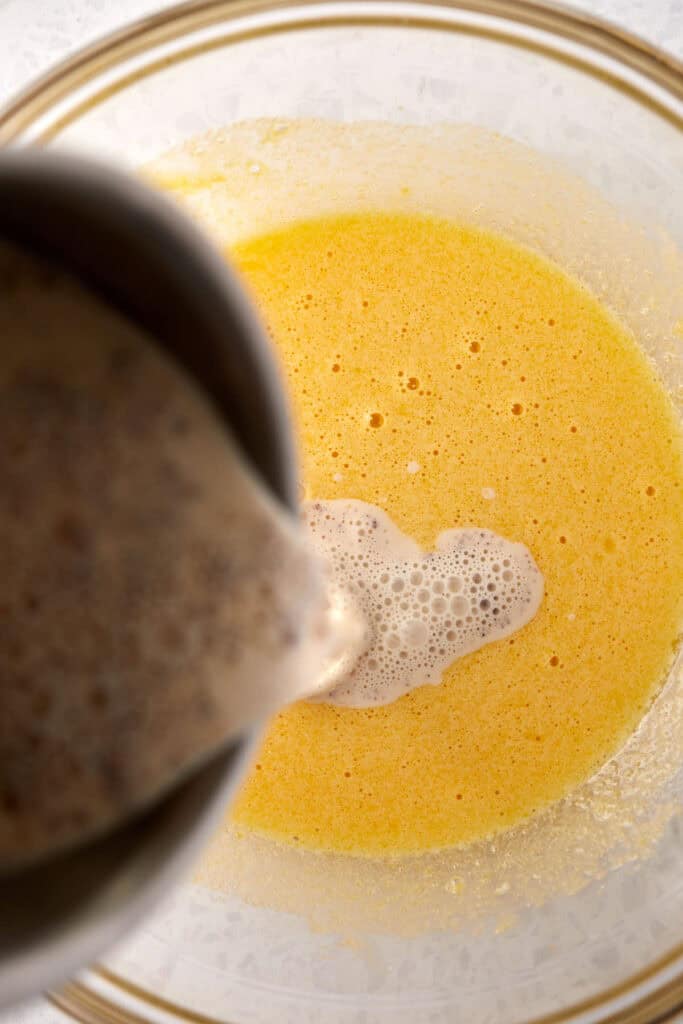 Batter being poured from a bowl into a larger bowl containing a yellow liquid mixture, creating bubbles on the surface. The yellow mixture suggests ingredients like eggs or butter.