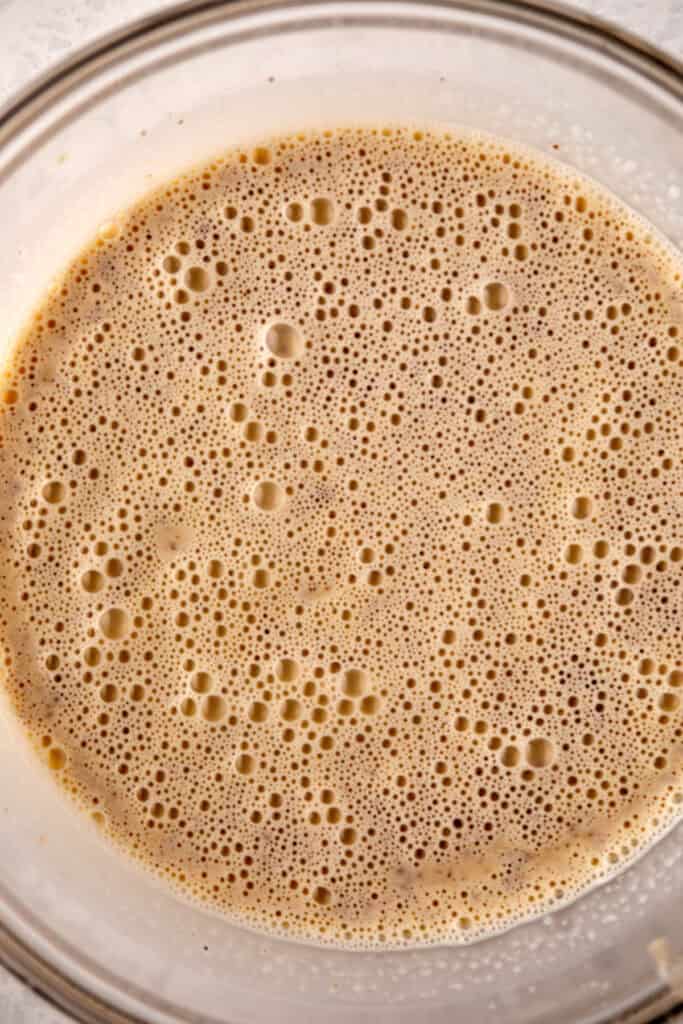 A glass bowl filled with a frothy, caramel-colored liquid mixture, showing numerous small bubbles on the surface. The texture suggests it could be a batter or a blended beverage.