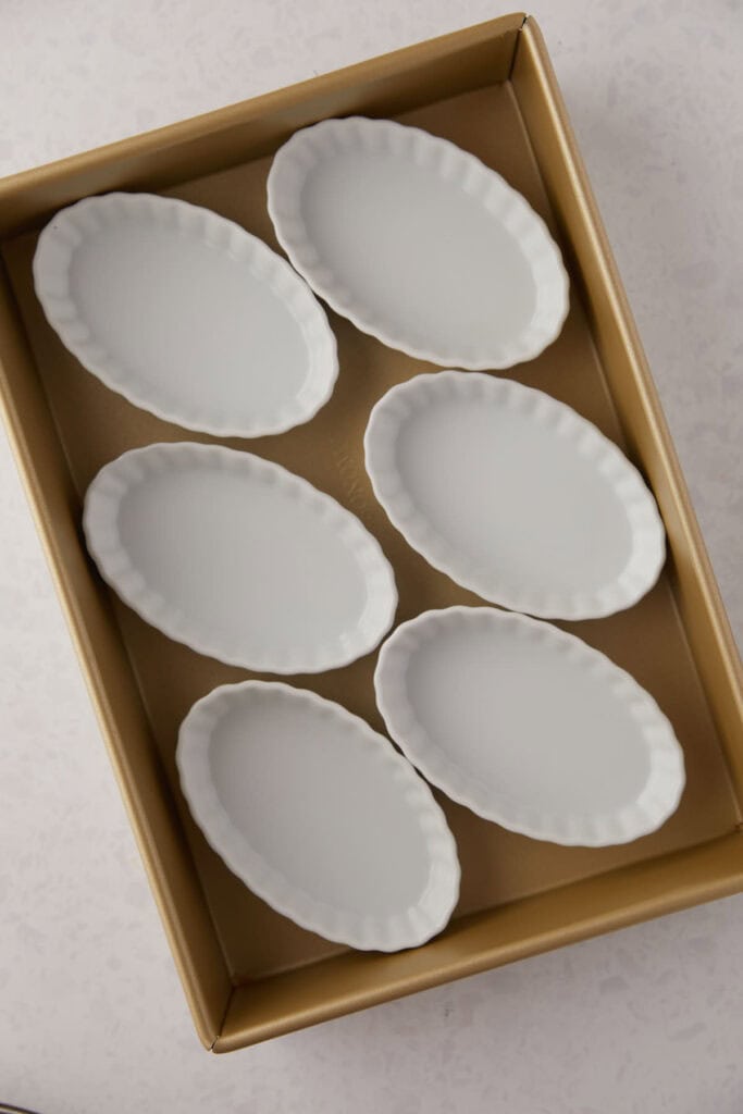 A box containing six white, oval-shaped ceramic plates with scalloped edges, neatly arranged in two columns of three on a light, textured surface.