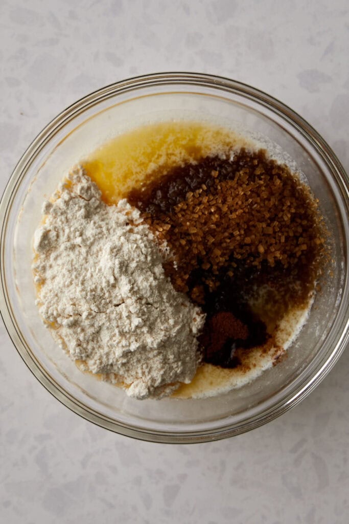 A glass bowl containing ingredients for baking: flour, turbinado sugar, melted butter, and cinnamon, all ready to be mixed. The bowl is placed on a light-colored countertop.