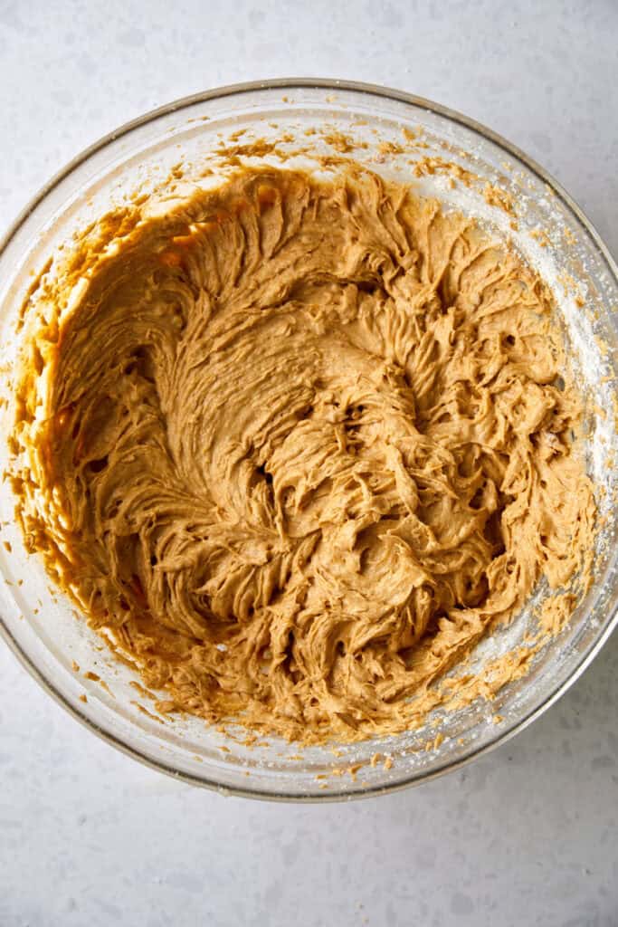 A glass bowl filled with creamy, light brown banana pumpkin batter sits on a white speckled countertop. The dough appears smooth and slightly swirled, ready for baking.