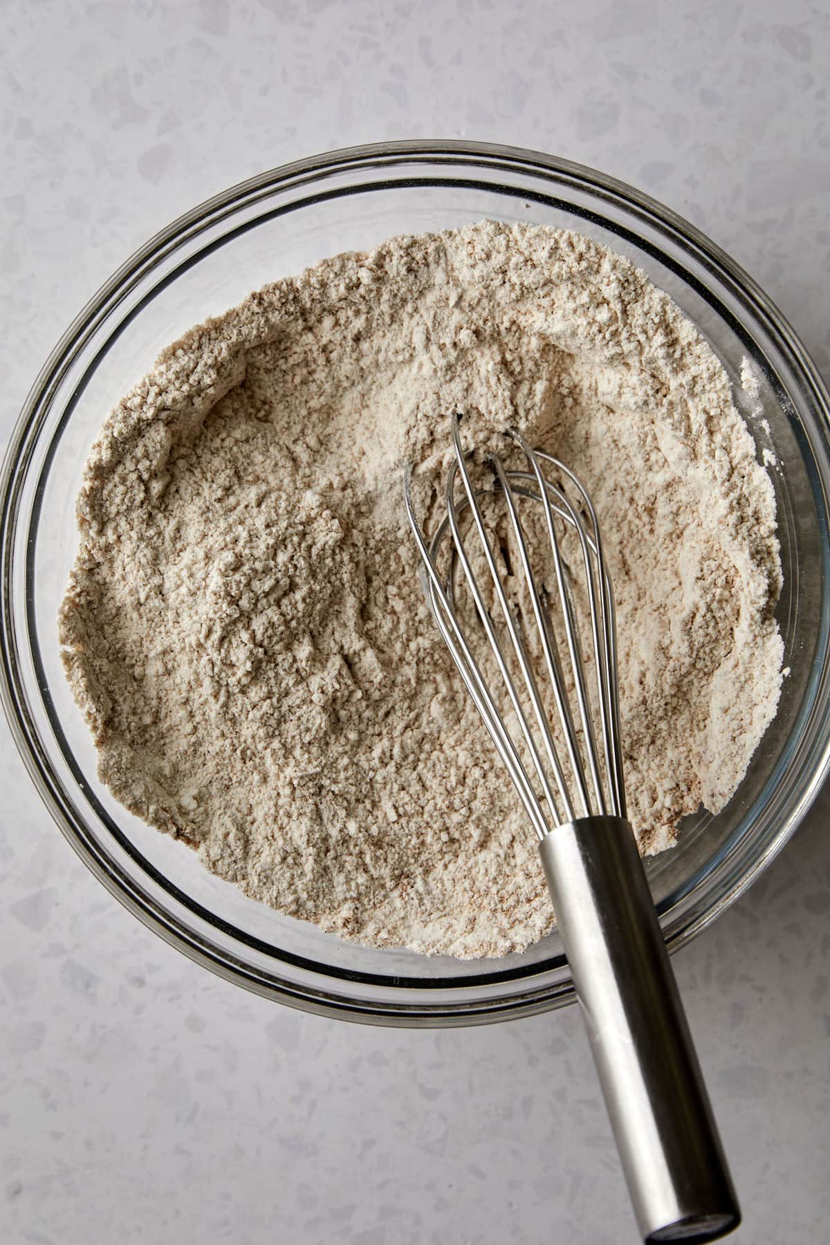 A glass bowl containing a mixture flour, baking powder, baking soda, salt and pumpkin pie spice is shown from above. A metal whisk rests inside the bowl, positioned to the right. The background is a light gray countertop.