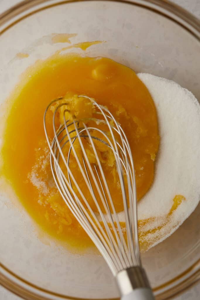 A glass bowl containing a mixture of bright yellow egg yolks and white sugar being whisked together by a metal whisk.