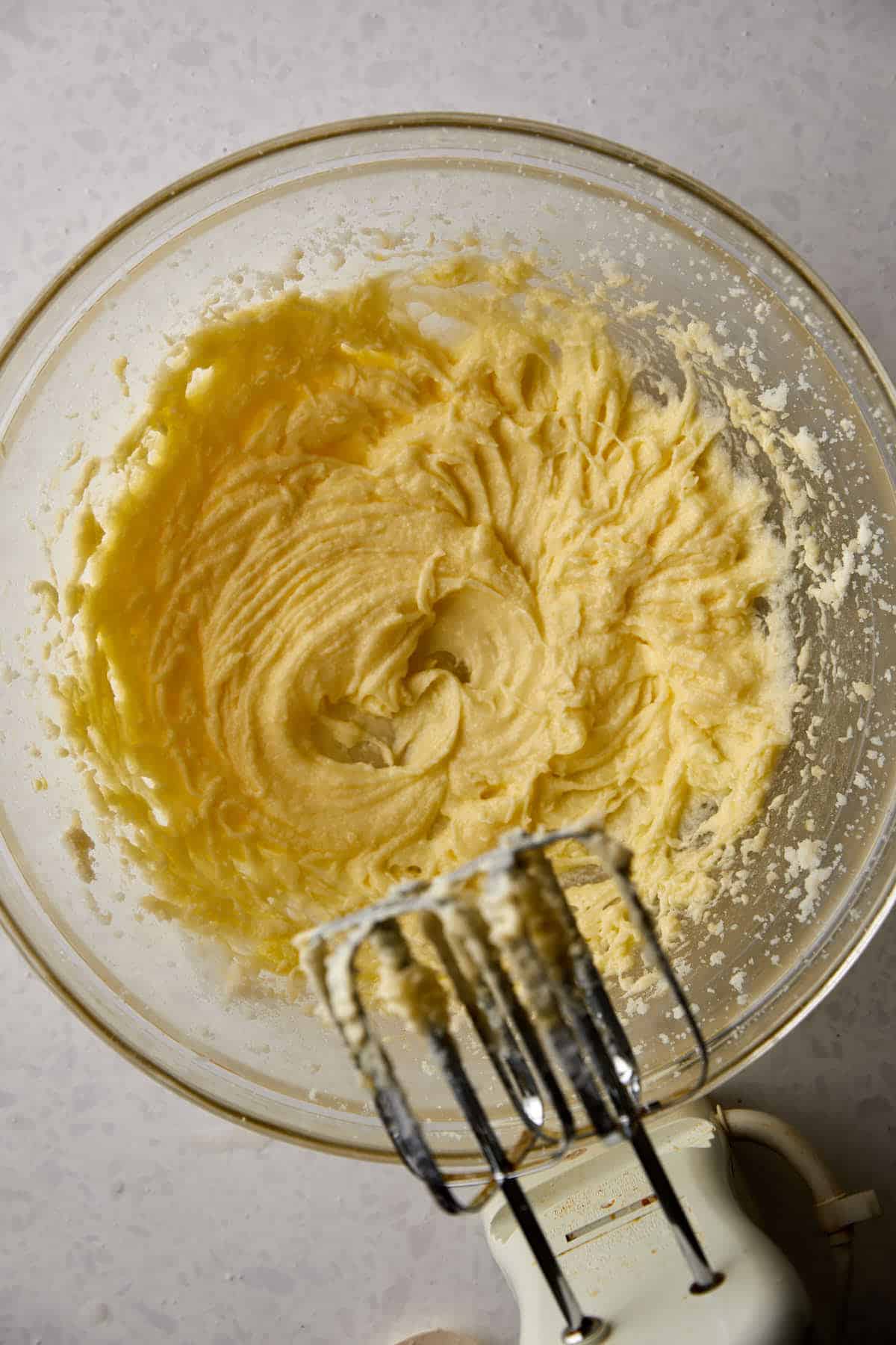 A glass bowl filled with creamy batter for moist and delicious Pumpkin Banana Muffins rests on a speckled countertop. An electric mixer, showcasing remnants of this easy fall recipe on its beaters, is partially visible in the foreground.