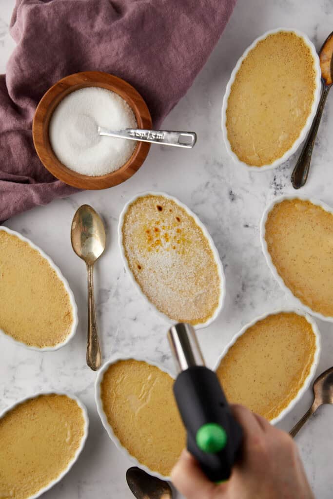 A person is using a kitchen torch to caramelize sugar on one of several oval dishes of crème brûlée. A bowl of sugar and a spoon are nearby on a marble surface. A purple cloth is partially visible.