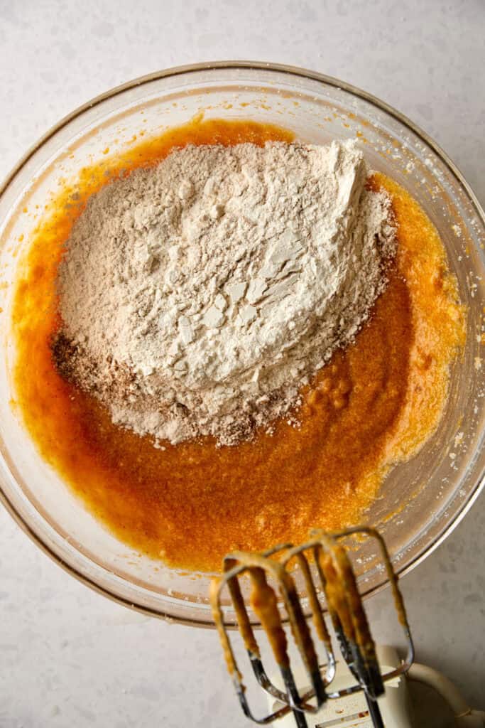 A mixing bowl with pumpkin banana muffin batter and flour mixture on top, sitting on a light countertop. An electric mixer with dough-covered beaters is positioned at the bottom right of the image.