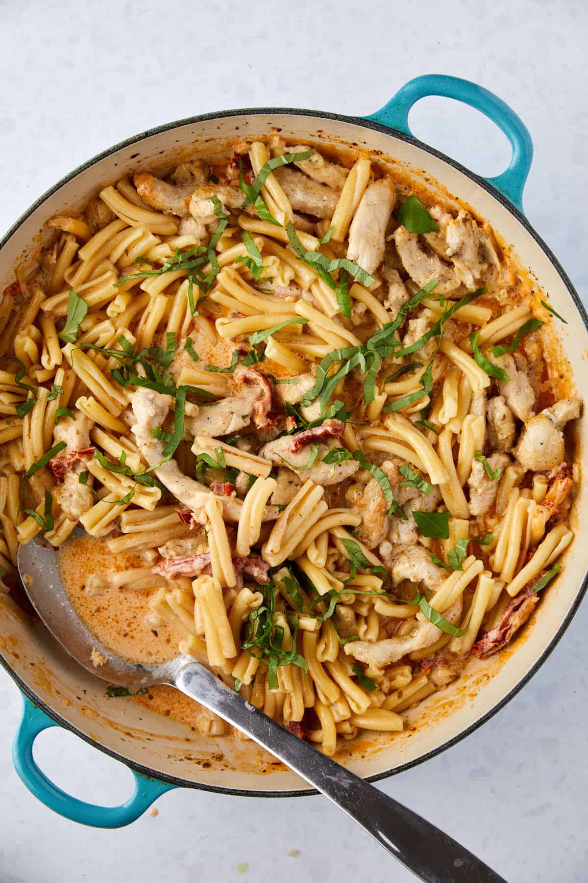 A top view of a blue pot filled with creamy pasta mixed with sliced chicken and sun-dried tomatoes. The dish is garnished with fresh basil leaves, and a spoon rests in the pot.