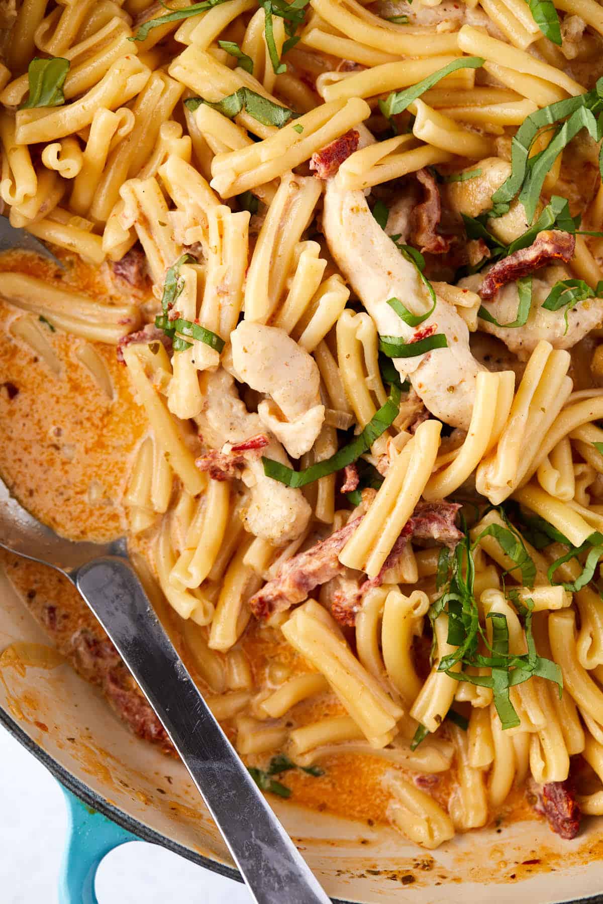 Close-up of a creamy pasta dish featuring short pasta, sliced chicken, sun-dried tomatoes, and a rich, orange-colored sauce. Garnished with fresh basil, served in a deep pan with a metal serving spoon.