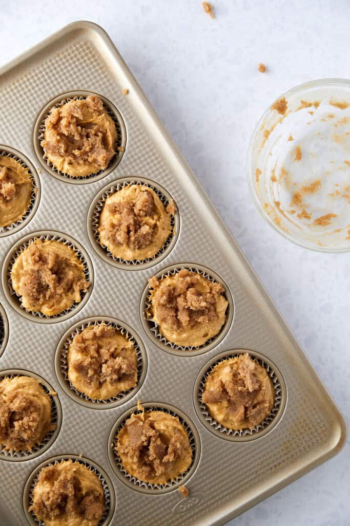 A baking tray with muffin liners filled with raw pumpkin banana batter, for muffins. The tray is on a light-colored surface, and a nearby bowl contains leftovers of the crumb sugar topping.