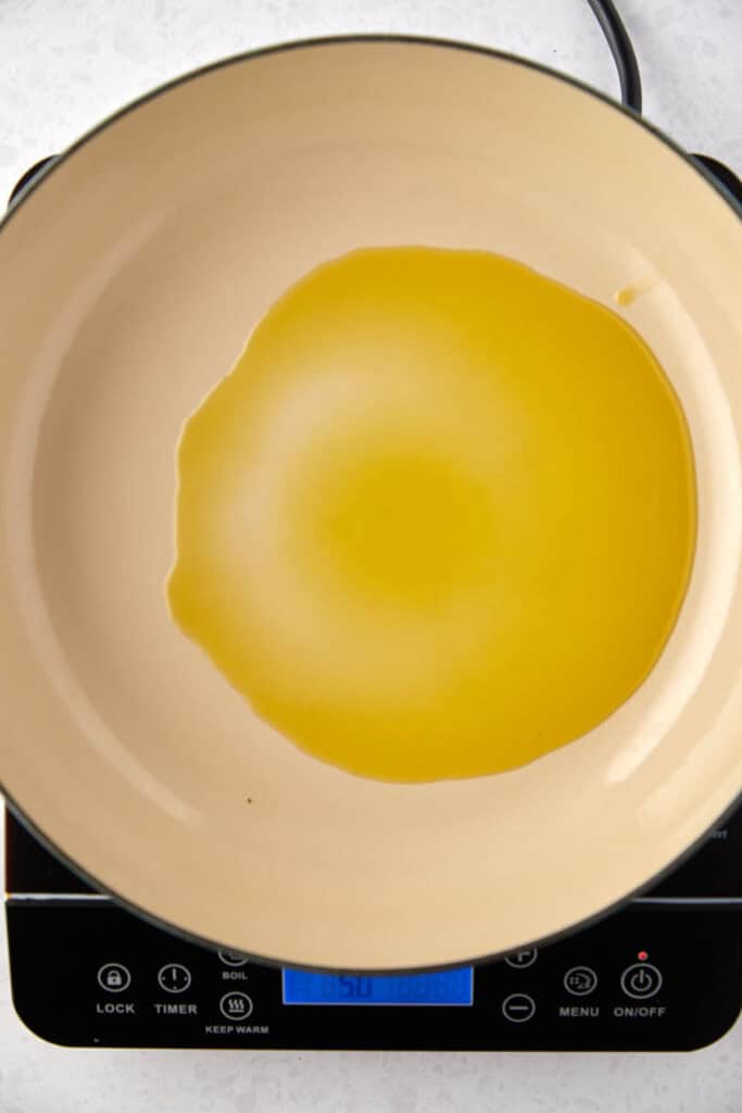 A top view of a beige pan on a black electric stove with a pool of olive oil spread evenly in the center. The stove's digital display shows numbers and symbols.