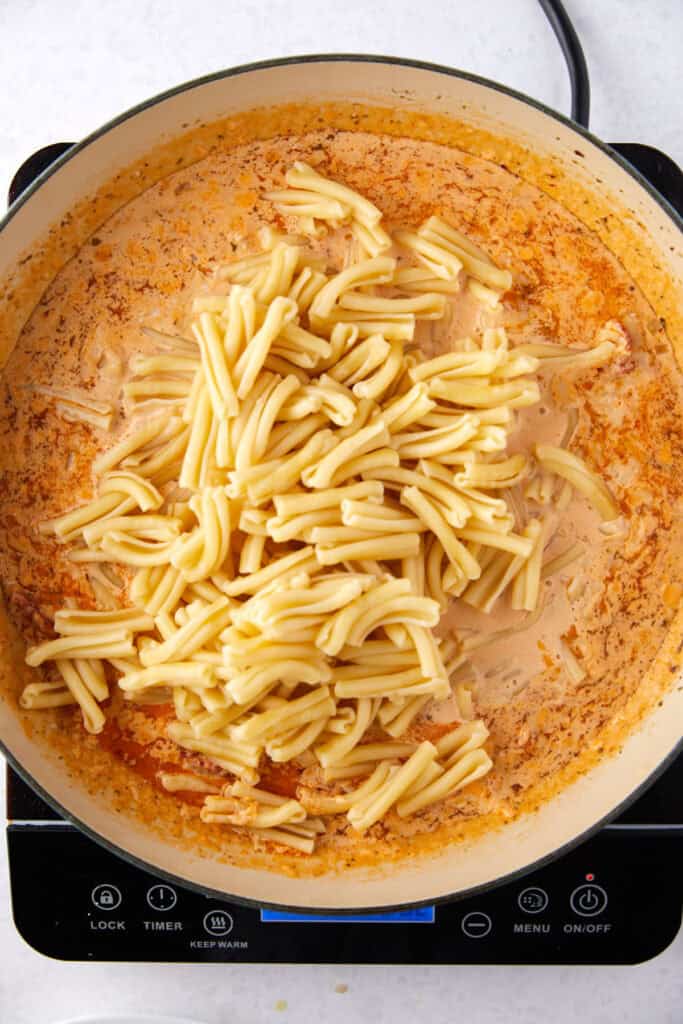 A pot with cooked pasta in a creamy, orange-red sauce, on an electric stove. The sauce is mixed with spices and the pasta is piled in the center. The stove displays various buttons and settings.