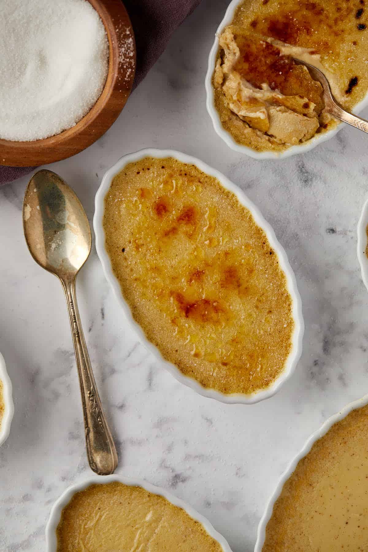 Oval ramekins filled with caramelized pumpkin crème brûlée are arranged on a marble surface. A spoon rests on the table next to a bowl of sugar. One ramekin is partially eaten, revealing the creamy interior.