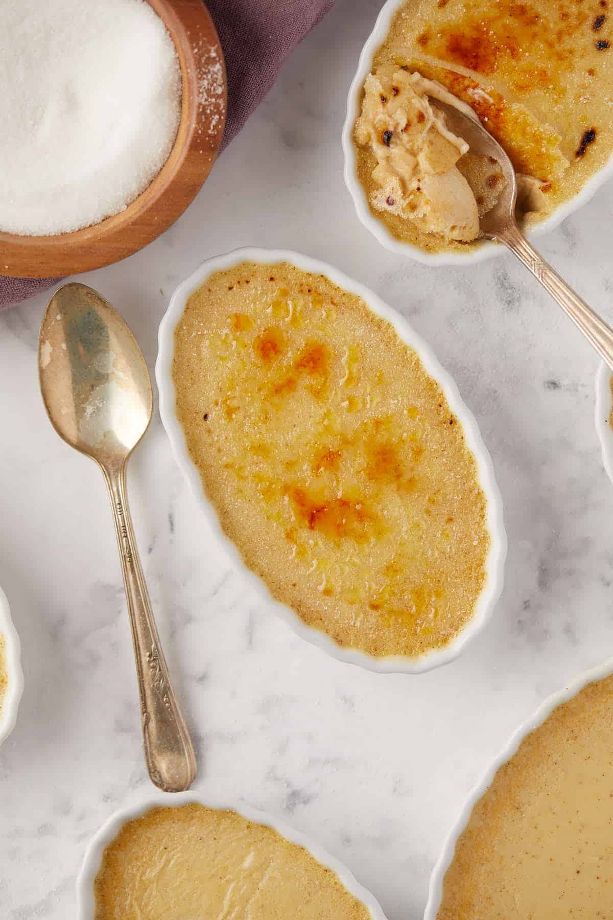 Oval dishes of crème brûlée with golden caramelized tops on a marble surface. A spoon rests on the surface, one indicating a bite taken. A wooden bowl of sugar is nearby.