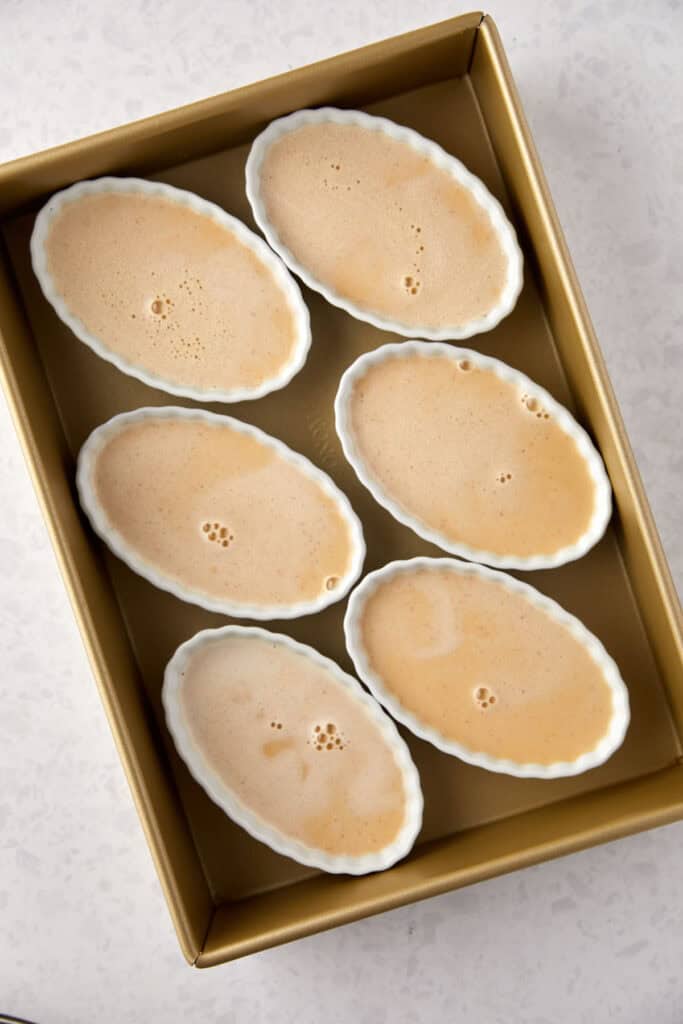 Six oval ramekins filled with a creamy, light orange batter are arranged in a gold baking tray on a white speckled countertop.