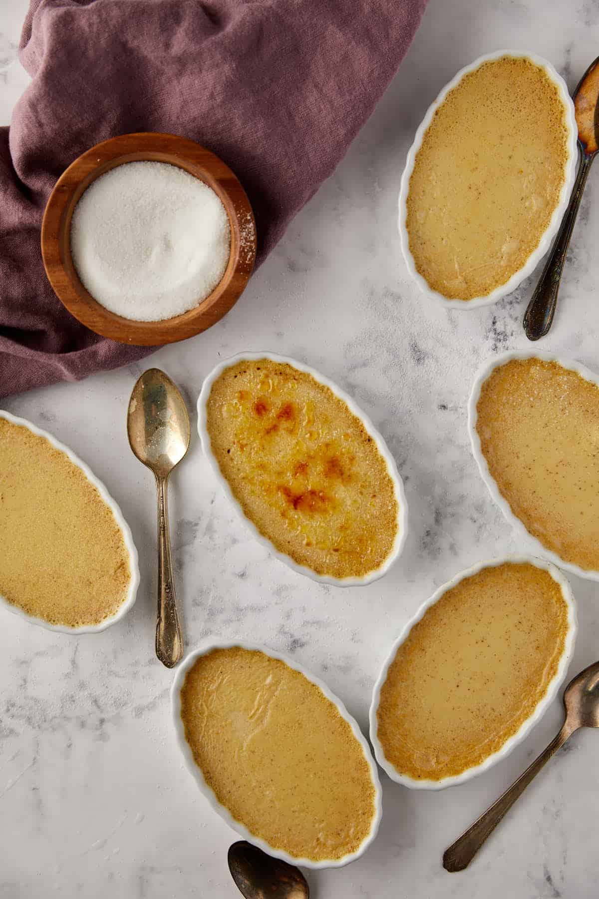 Oval ramekins of crème brûlée sit on a marble surface. A few have caramelized tops. Several spoons are placed beside them. A wooden bowl filled with sugar is next to a purple cloth in the background.