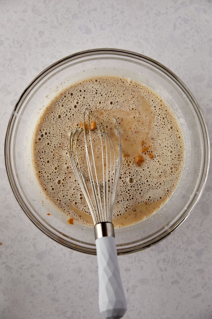 A glass bowl with a mixture of wet ingredients and chunks of brown sugar being whisked. The mixture is light brown and bubbly. A white-handled whisk rests inside the bowl on a speckled light gray countertop.