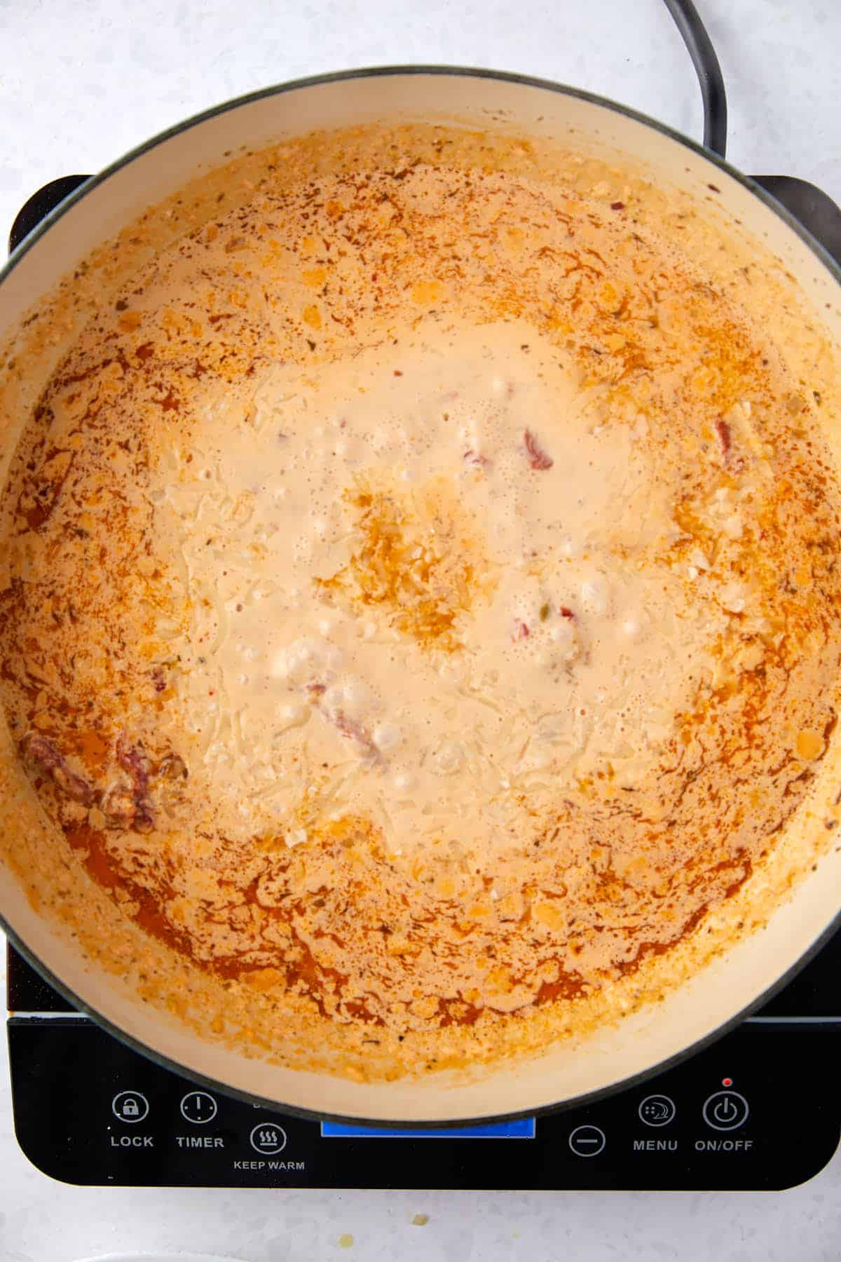 A pot of cream with spices simmers on an electric stovetop, displaying a rich orange hue with bits of sun dried tomatoes.